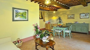 A dining area with a wooden table and chairs, a kitchen in the background, and a stone wall. - Estate in Monte Serra with Pool & Olive Views (Pisa)