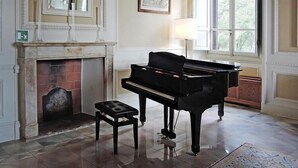 Un piano à queue dans une pièce spacieuse, dotée d’une cheminée, d’une grande fenêtre et d’un tableau encadré au mur.