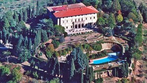Une vue aérienne d’un grand bâtiment rectangulaire, doté d’un toit rouge, entouré d’un jardin aménagé avec des arbres et une piscine.