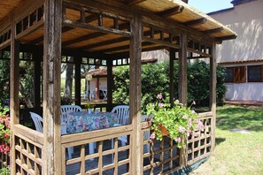 A wooden gazebo with a table and chairs, surrounded by a wooden fence and potted plants.