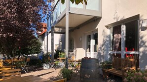 An outdoor seating area with wooden tables and benches, surrounded by potted plants and a large barrel. - TH39 Trend Hotel (Bad Vilbel)
