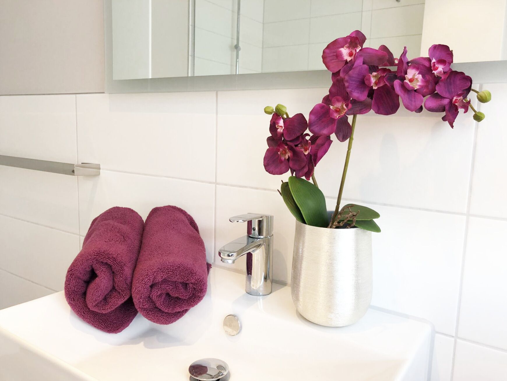 Un baño con lavamanos blanco, espejo y orquídea en maceta.