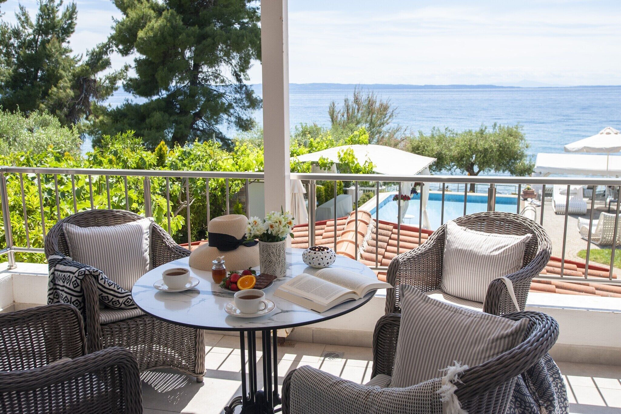 Un balcone con mobili in vimini, un tavolo con un libro, una tazza e un piatto di cibo, con vista su una piscina e uno specchio d'acqua.