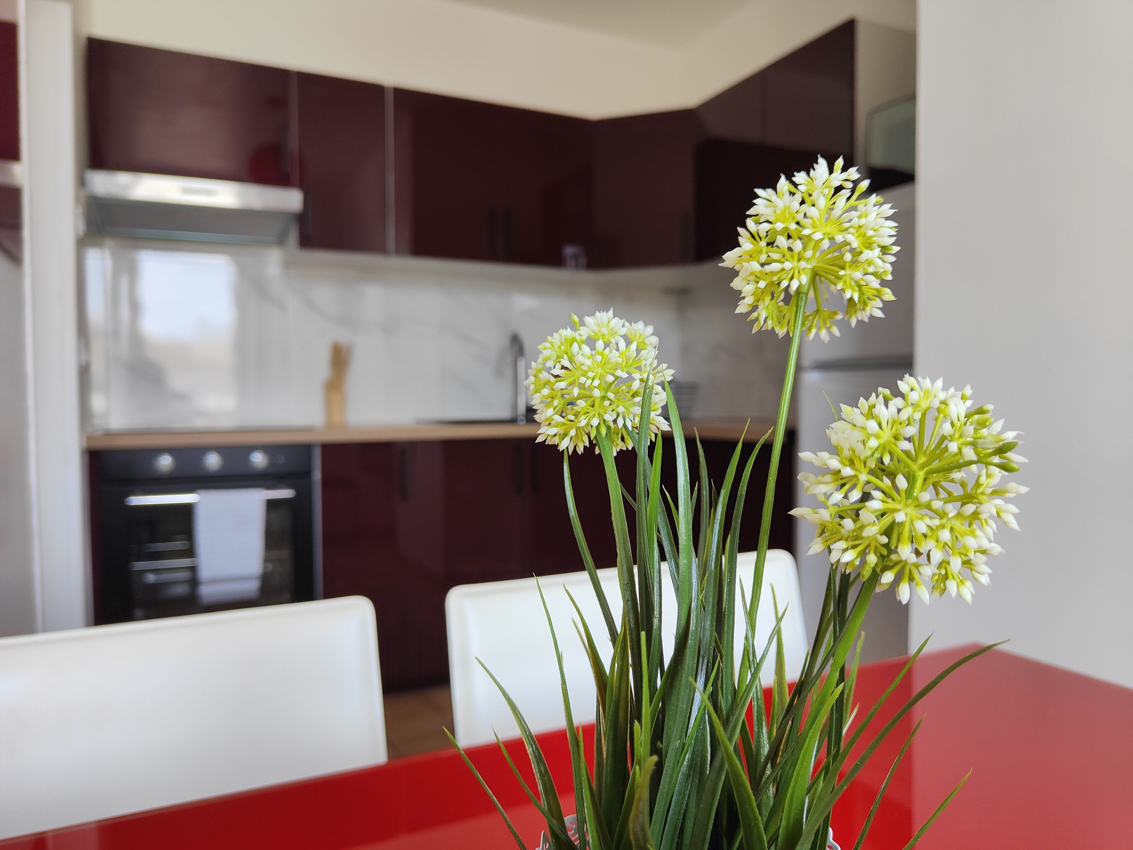 A modern kitchen with dark cabinets, a stainless steel range hood, and a red dining table with a vase of flowers.