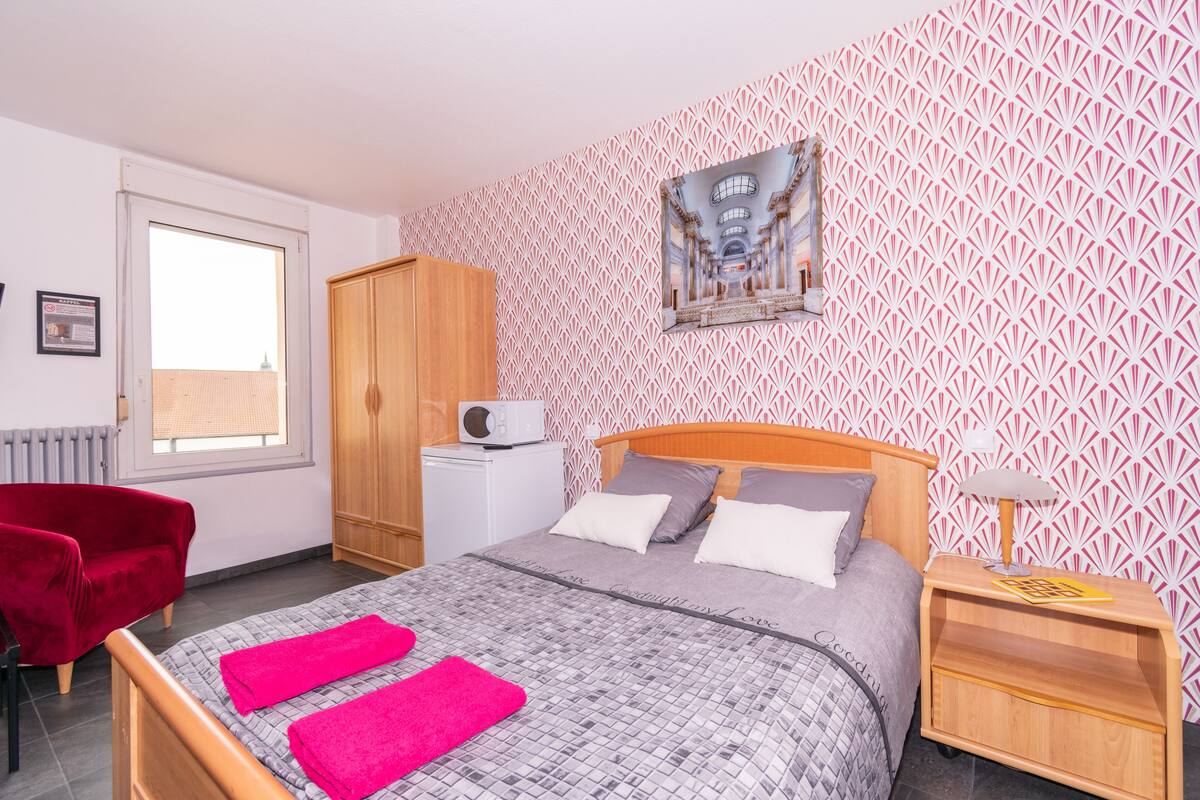 Een slaapkamer met een bed, een houten garderobe, een rode stoel, een nachtkastje en een foto aan de muur.