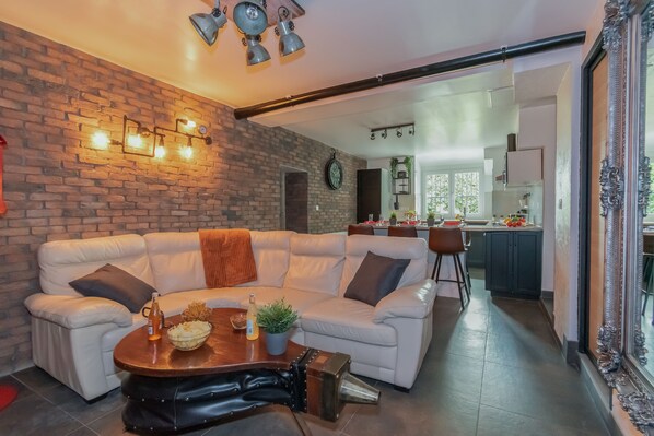 A modern living room with a brick wall, a white leather sofa, a wooden coffee table, and a kitchen in the background.