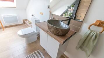 Un baño moderno con un inodoro blanco, un tocador de madera con un lavabo grande, un perchero y un espejo.
