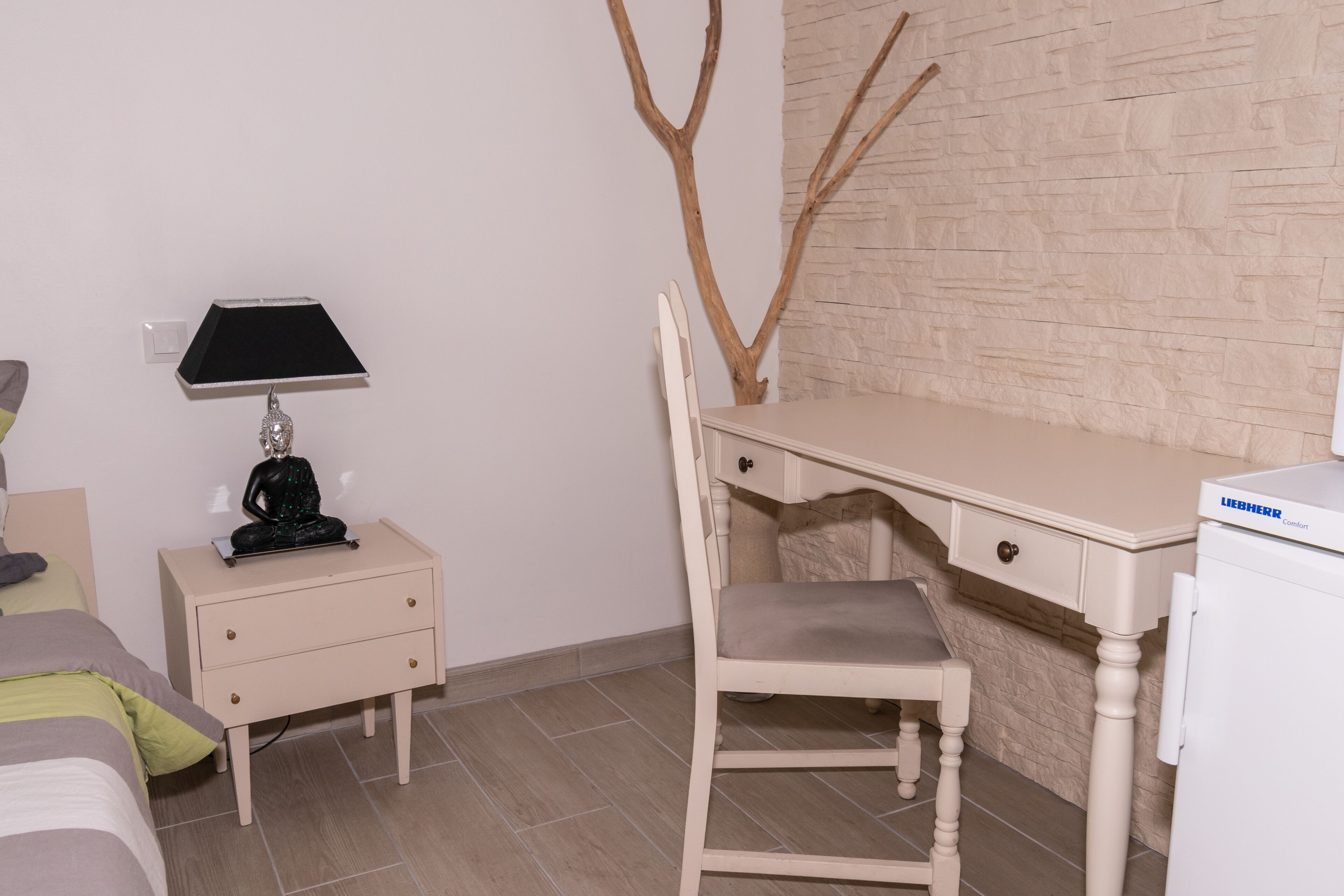 A bedroom with a bed, a nightstand with a lamp, a desk with a chair, and a white wardrobe.