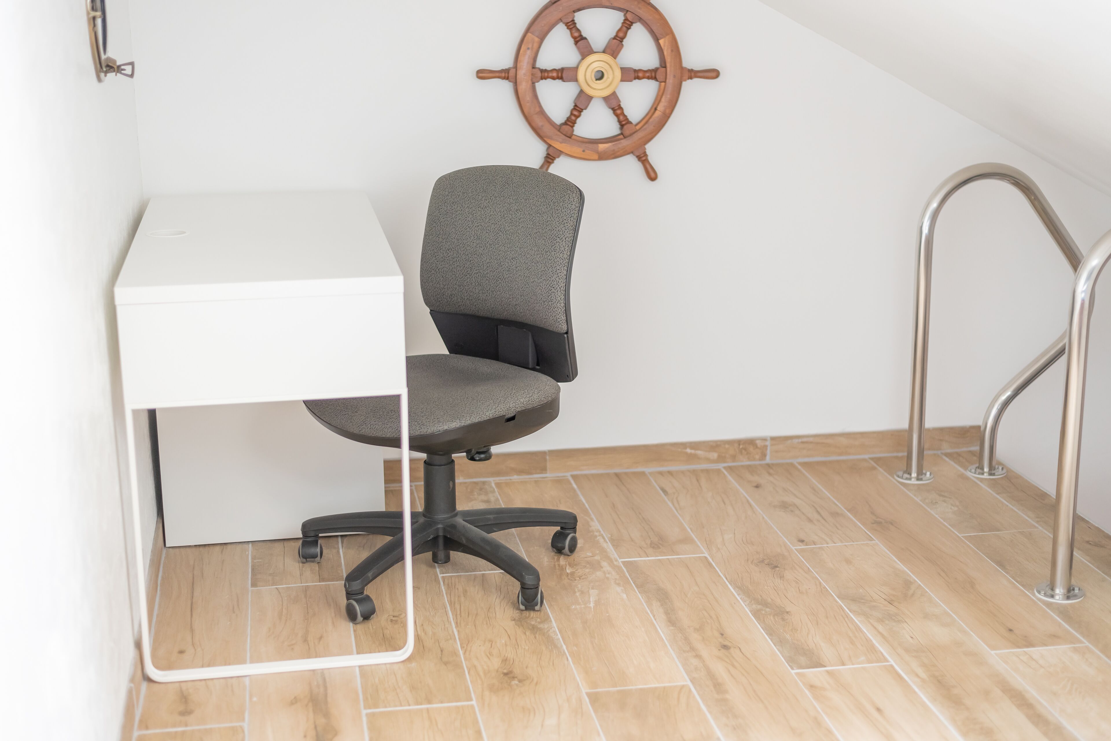 Un pequeño espacio de oficina con un escritorio blanco, una silla de oficina gris, una rueda de barco de madera en la pared y una escalera de metal.