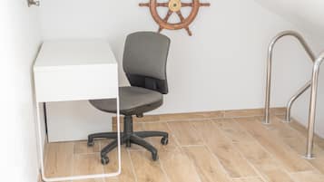 Un pequeño espacio de oficina con un escritorio blanco, una silla de oficina gris, una rueda de barco de madera en la pared y una escalera de metal.