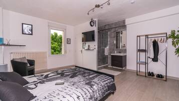 A modern bedroom with a large bed, a walk-in closet, a shower area, and a view of greenery outside.
