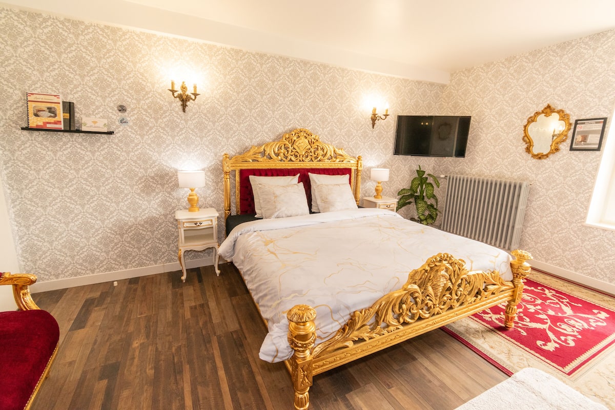 Een luxueuze slaapkamer met een groot bed, gouden accenten, een tapijt met een rood-wit patroon, een klein nachtkastje met een lamp en een aan de muur bevestigde televisie.
