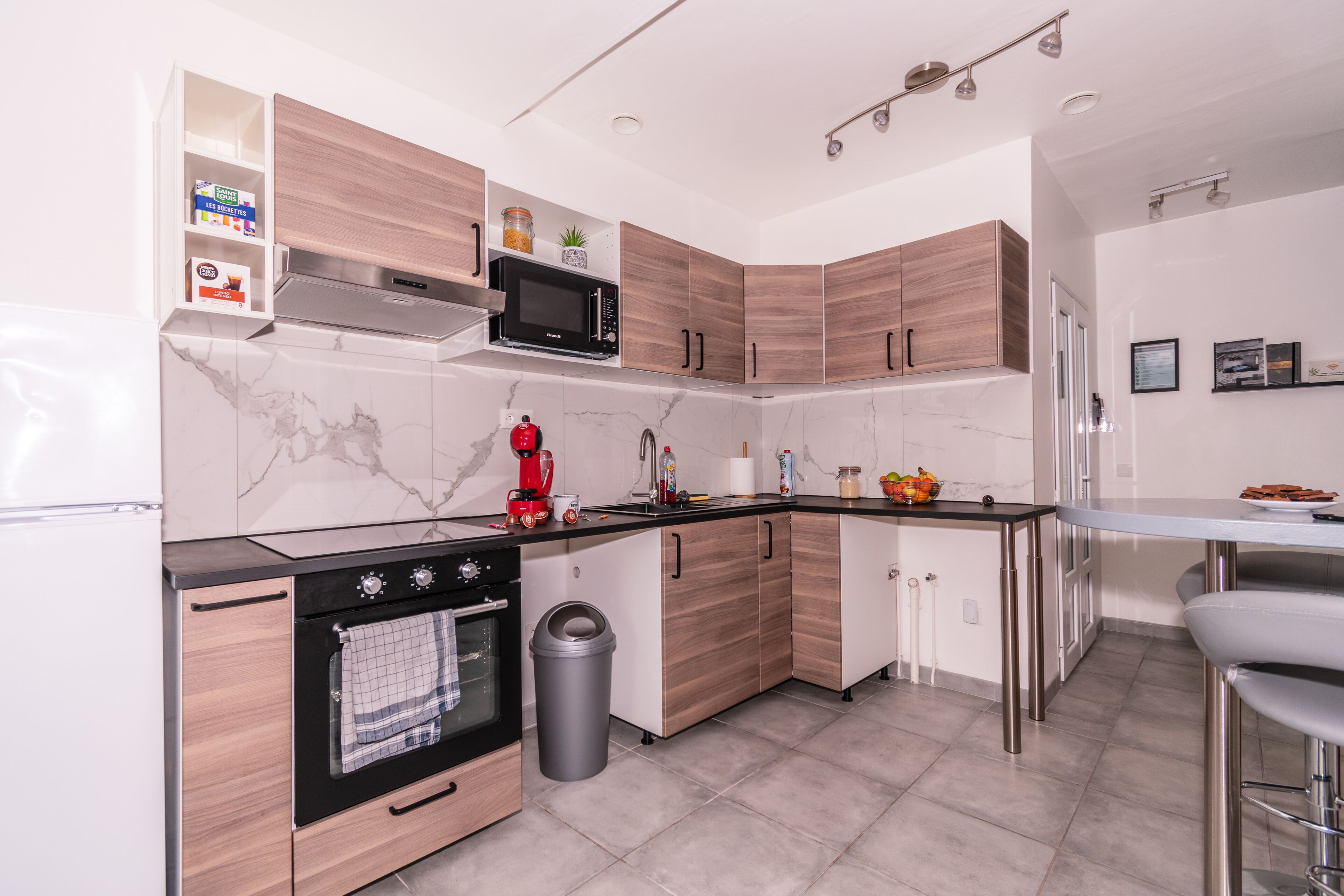 A modern kitchen with wooden cabinets, a black microwave, a coffee maker, and a trash can.