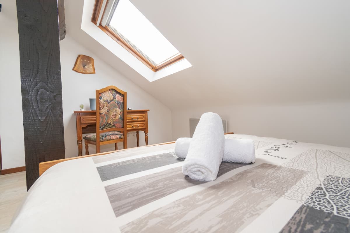 Een slaapkamer met een dakraam, een bed met een decoratieve spreidek, een houten dressoir en een stoel.