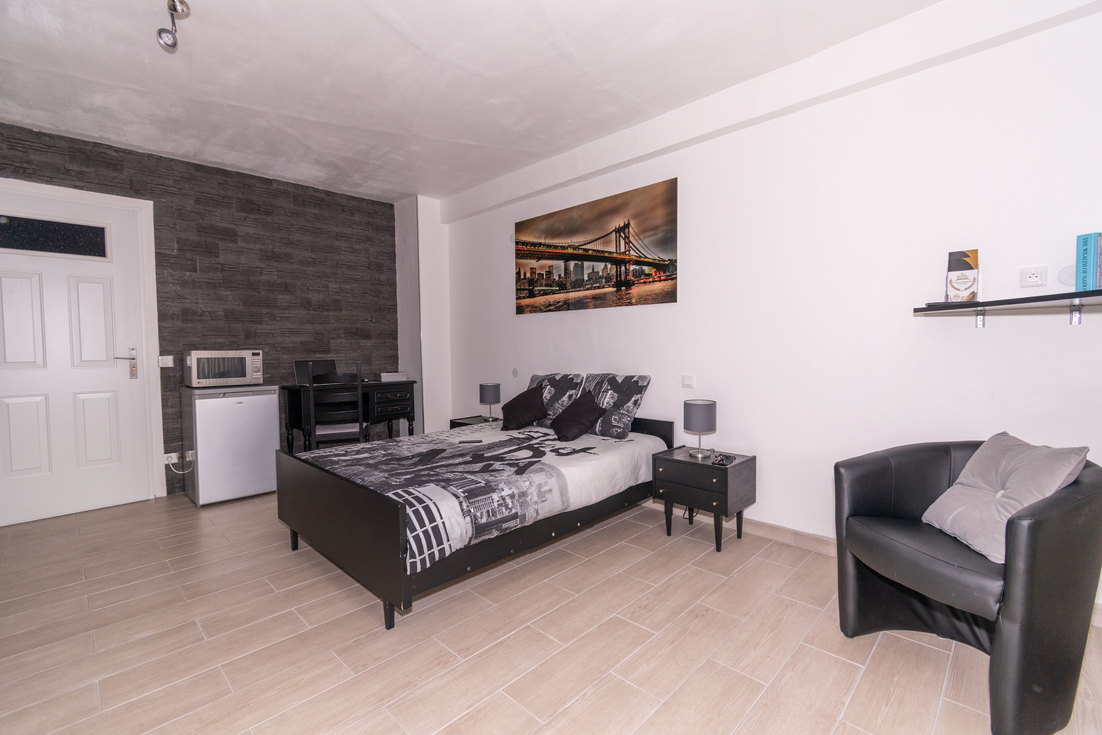 Un dormitorio con una cama, un sillón de cuero negro, un escritorio pequeño, un microondas y un cuadro de un puente.