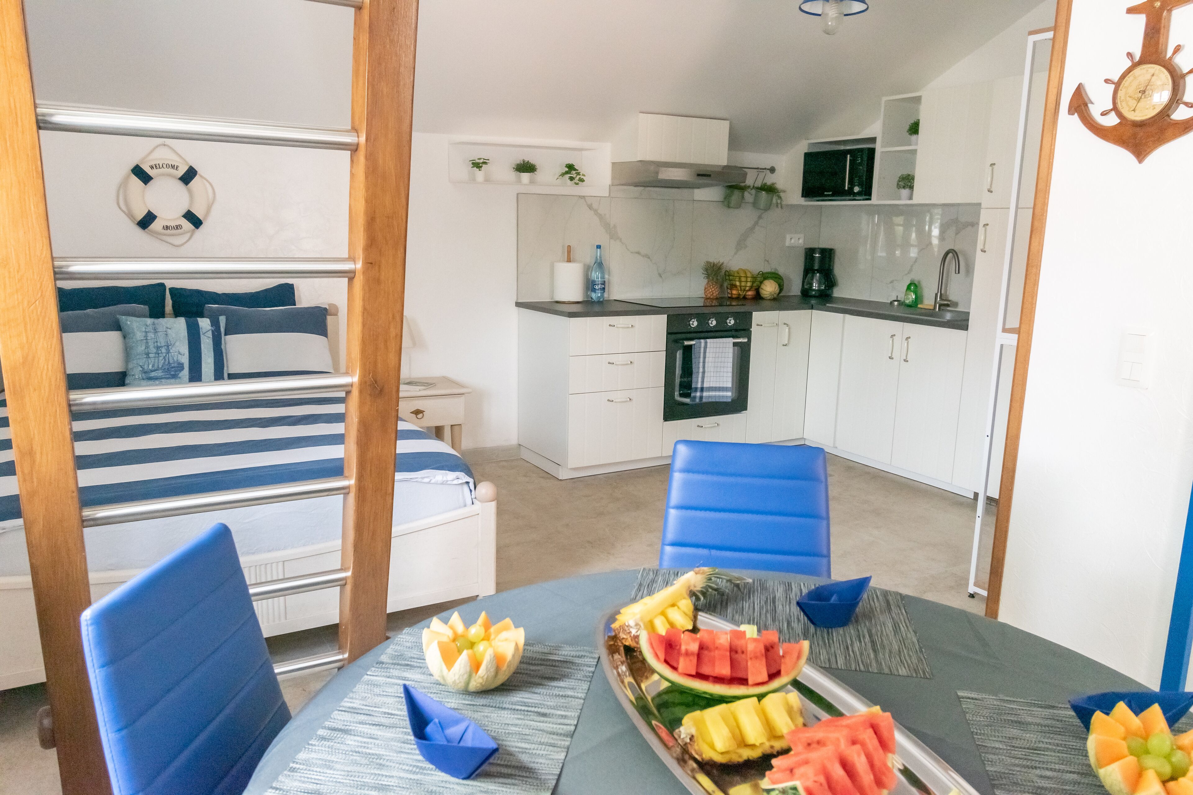 Une petite pièce comprenant un lit, une table à manger avec des fruits, un coin cuisine et une chaise bleue.