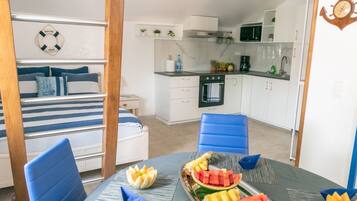 Une petite pièce comprenant un lit, une table à manger avec des fruits, un coin cuisine et une chaise bleue.
