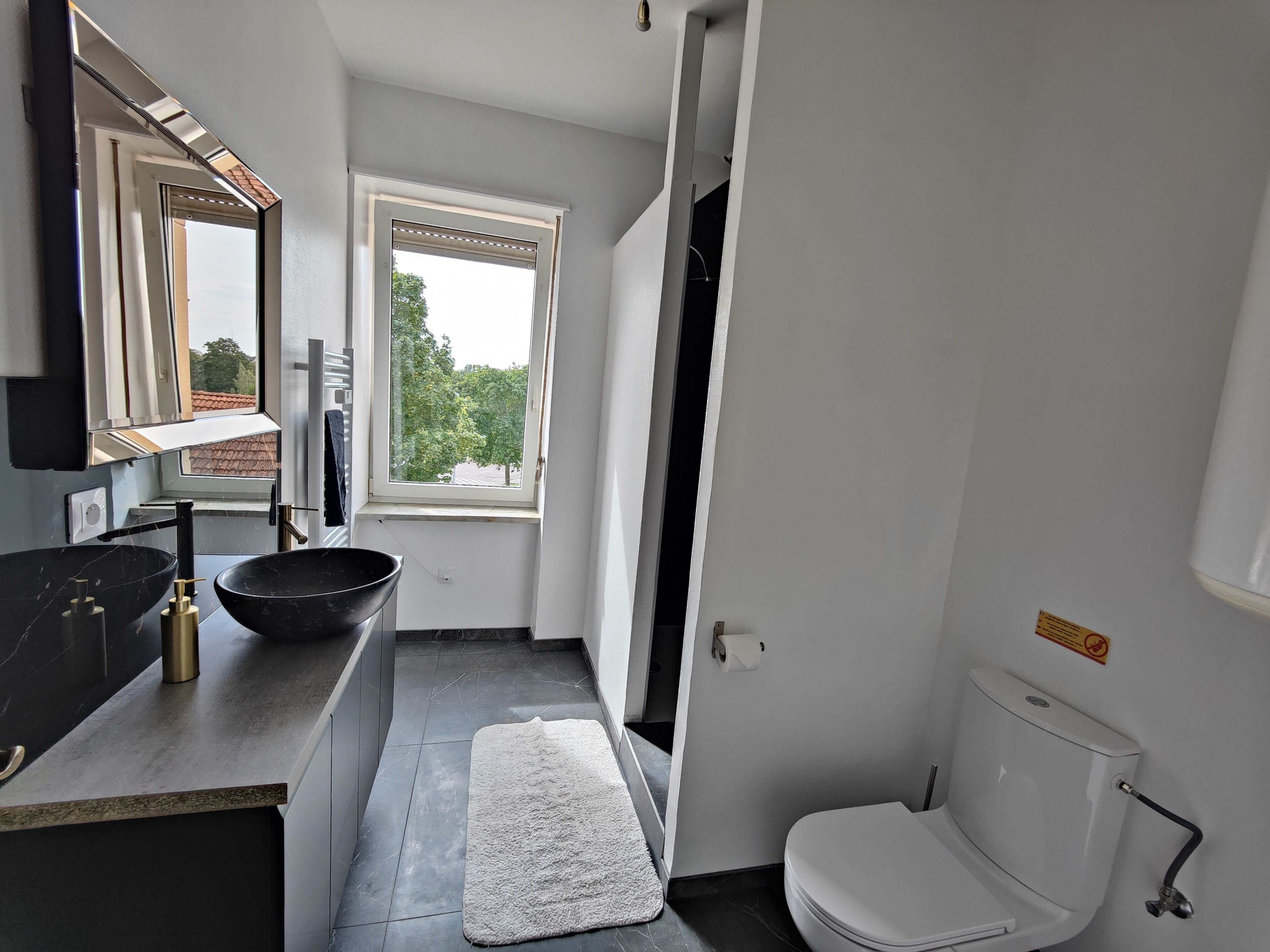 Ein modernes Badezimmer mit einem schwarzen Waschbecken, einer weißen Toilette und einem Fenster mit Blick auf Bäume.