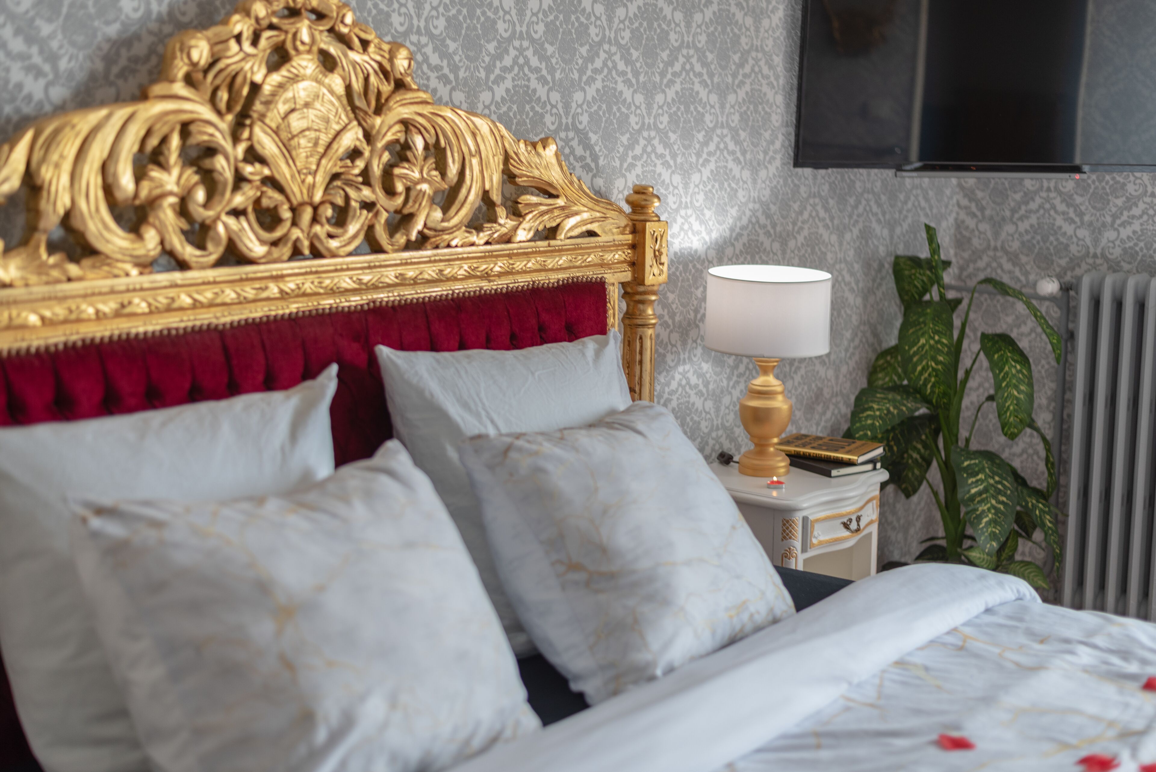Een bed met een gouden hoofdeinde, witte en rode kussens, een nachtkastje met een lamp en een plant.