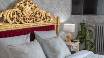 Een bed met een gouden hoofdeinde, witte en rode kussens, een nachtkastje met een lamp en een plant.