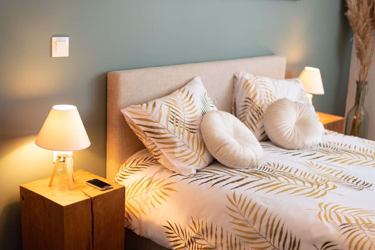 Een net opgemaakt bed met een sprei met bladmotief, twee bedlampjes en een smartphone op een nachtkastje van hout.
