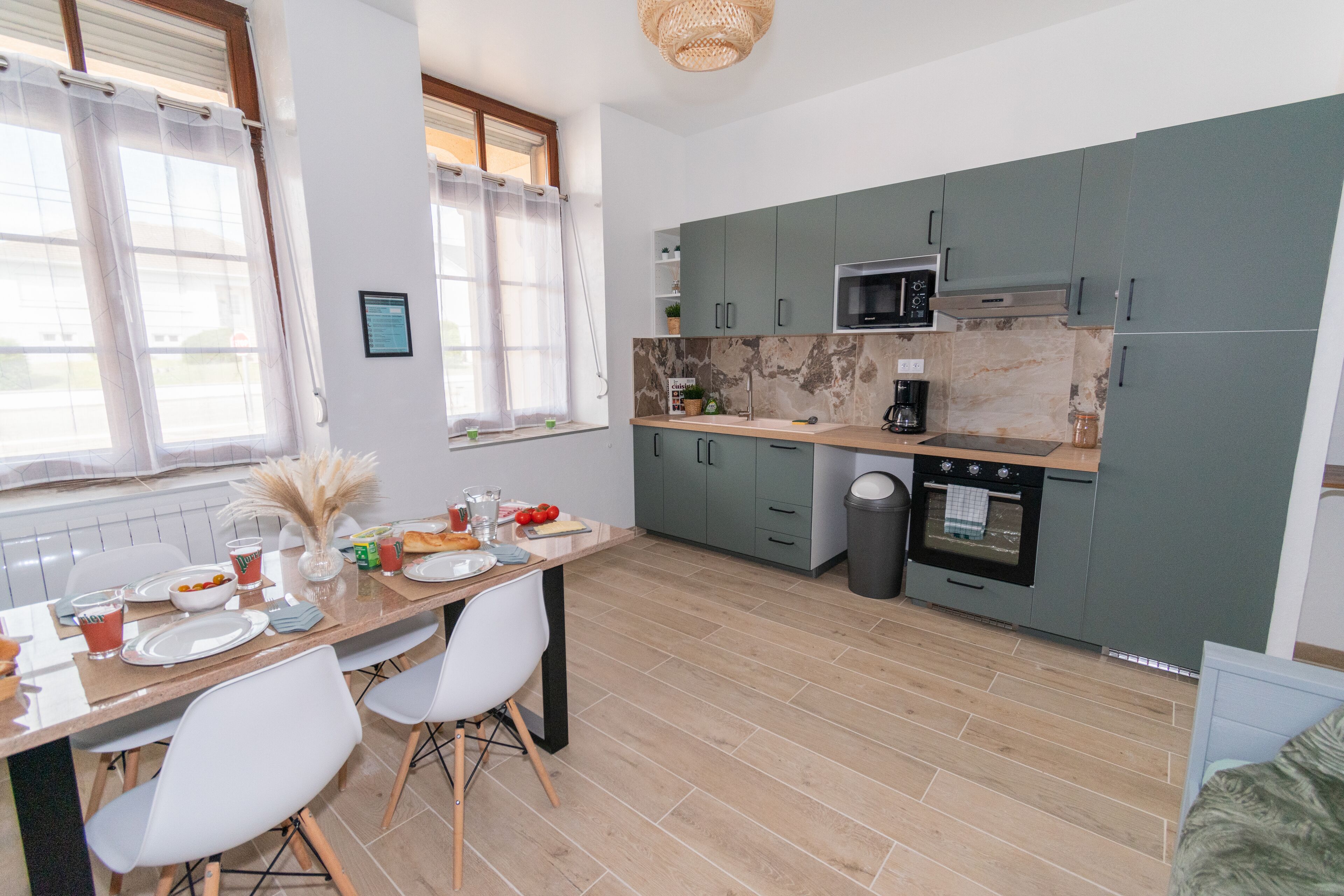 Eine moderne Küche mit grauen Schränken, einer Mikrowelle und einer Kaffeemaschine. Es gibt einen Esstisch mit Stühlen und ein Fenster mit Vorhängen.