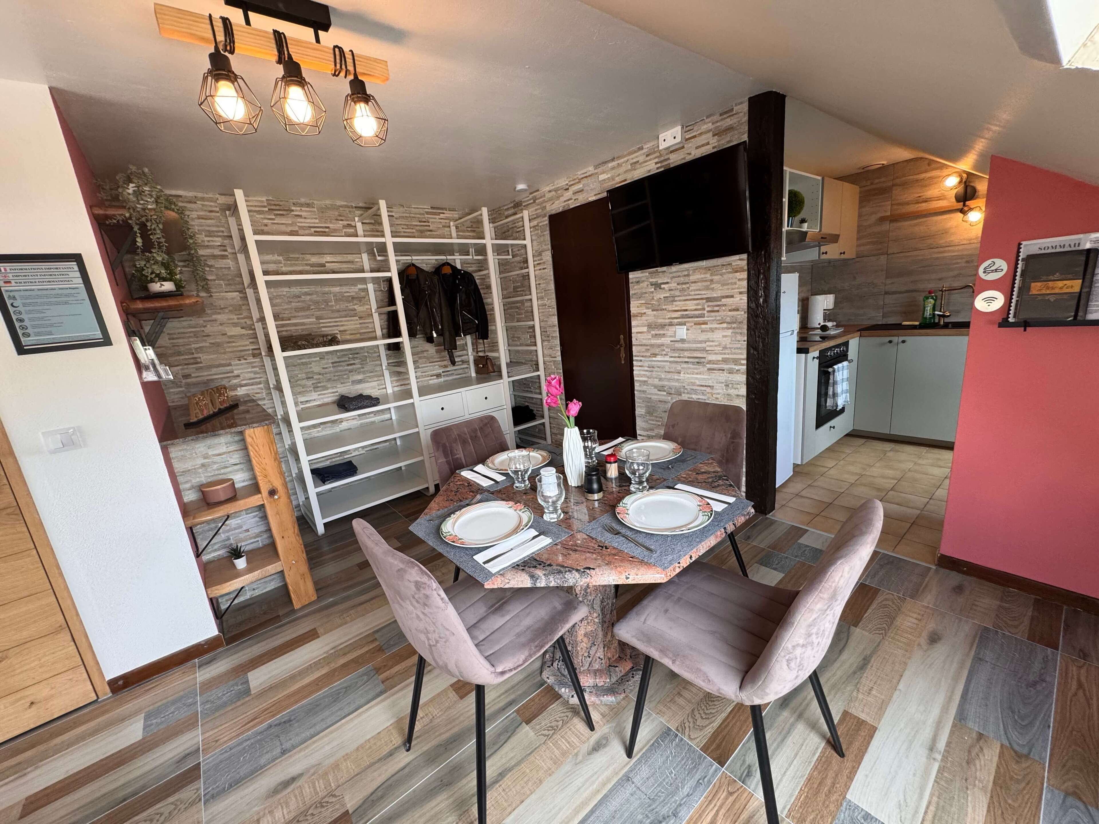 A dining area with a wooden table set for four, surrounded by cushioned chairs. A white cabinet with a glass top is visible to the left.