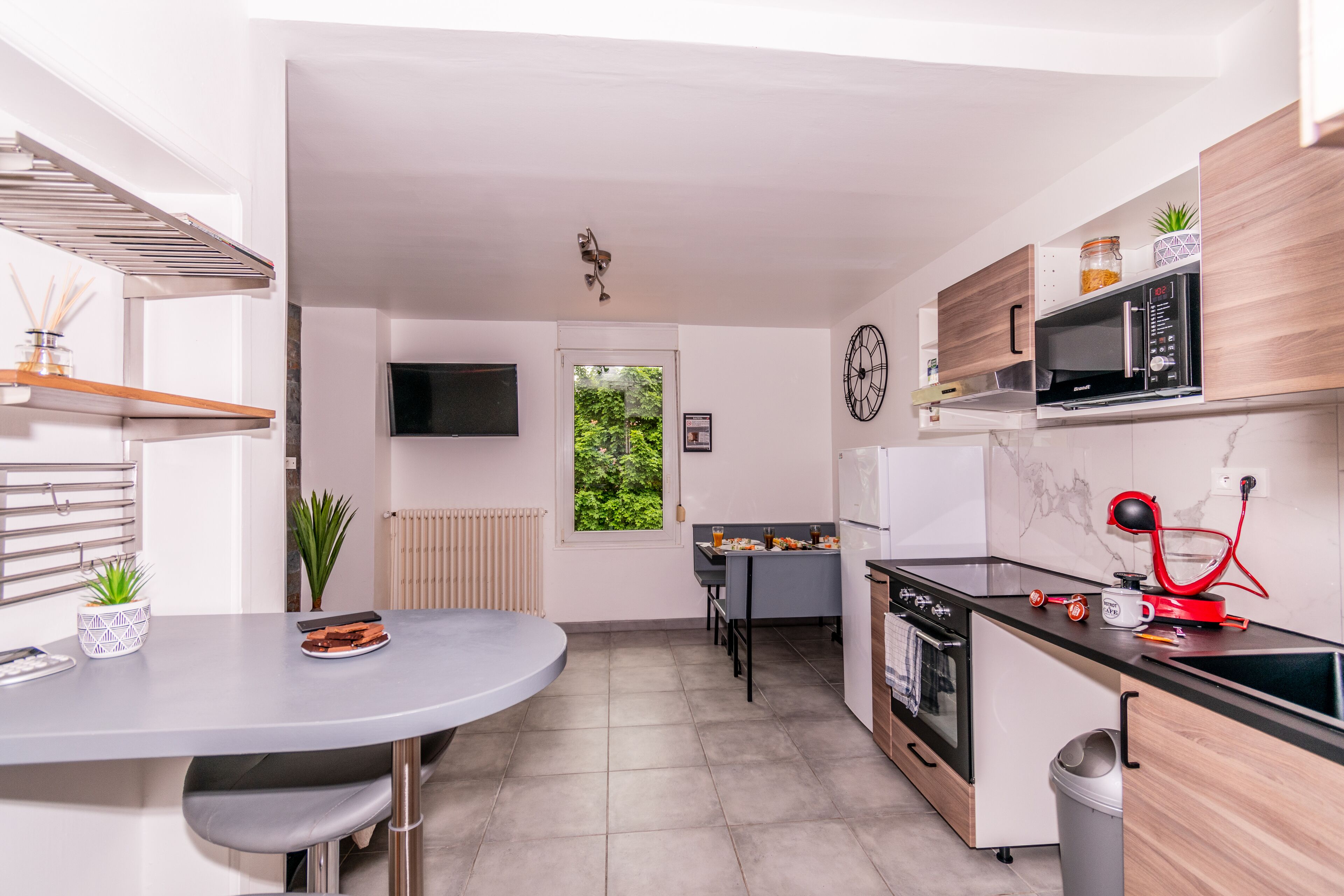 Una cuina moderna amb zona de menjador, televisió penjada a la paret i finestra amb vista a vegetació.