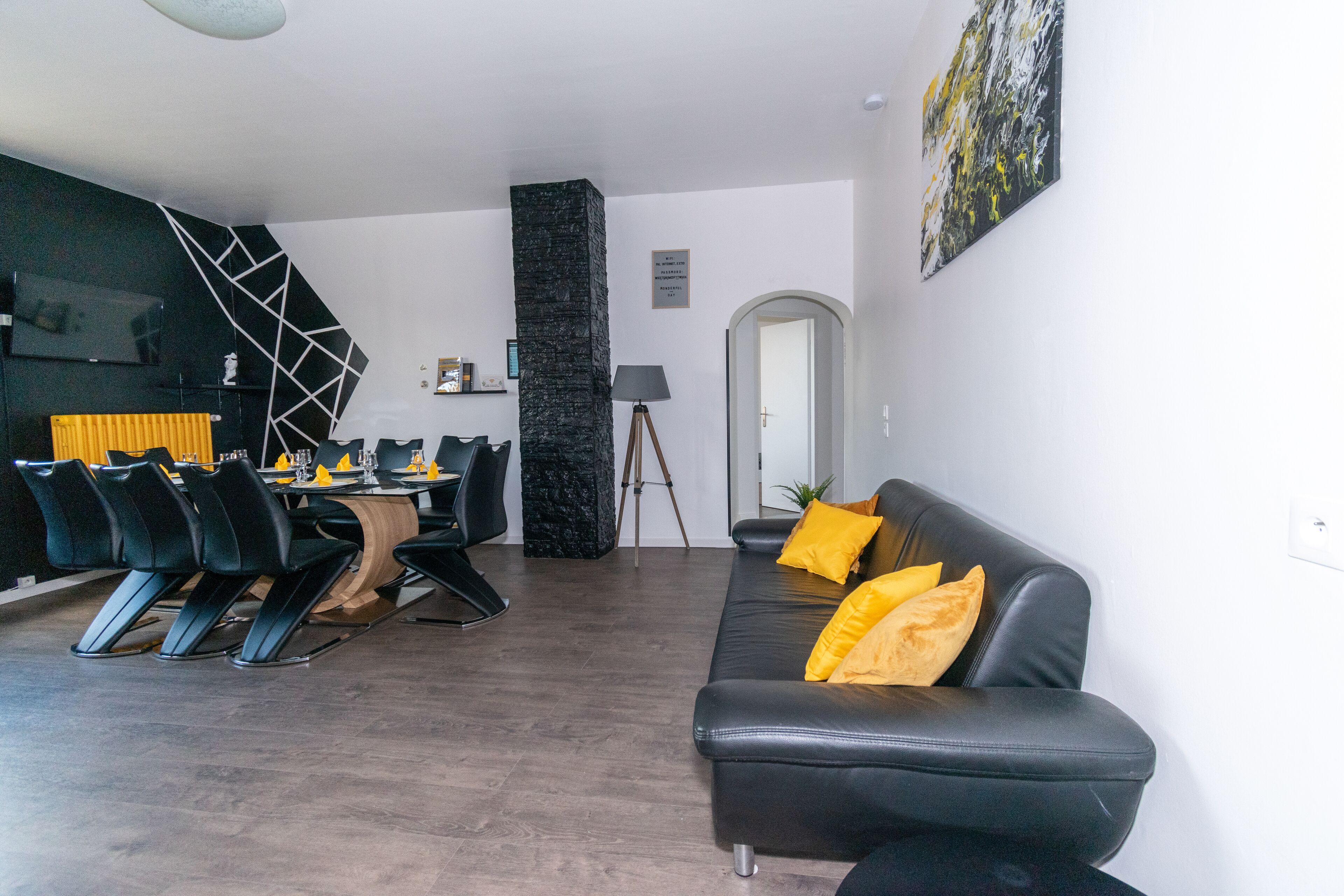 A modern living room with a black leather sofa, dining table, and chairs. There is a black accent wall with a geometric pattern and a tall black pillar.