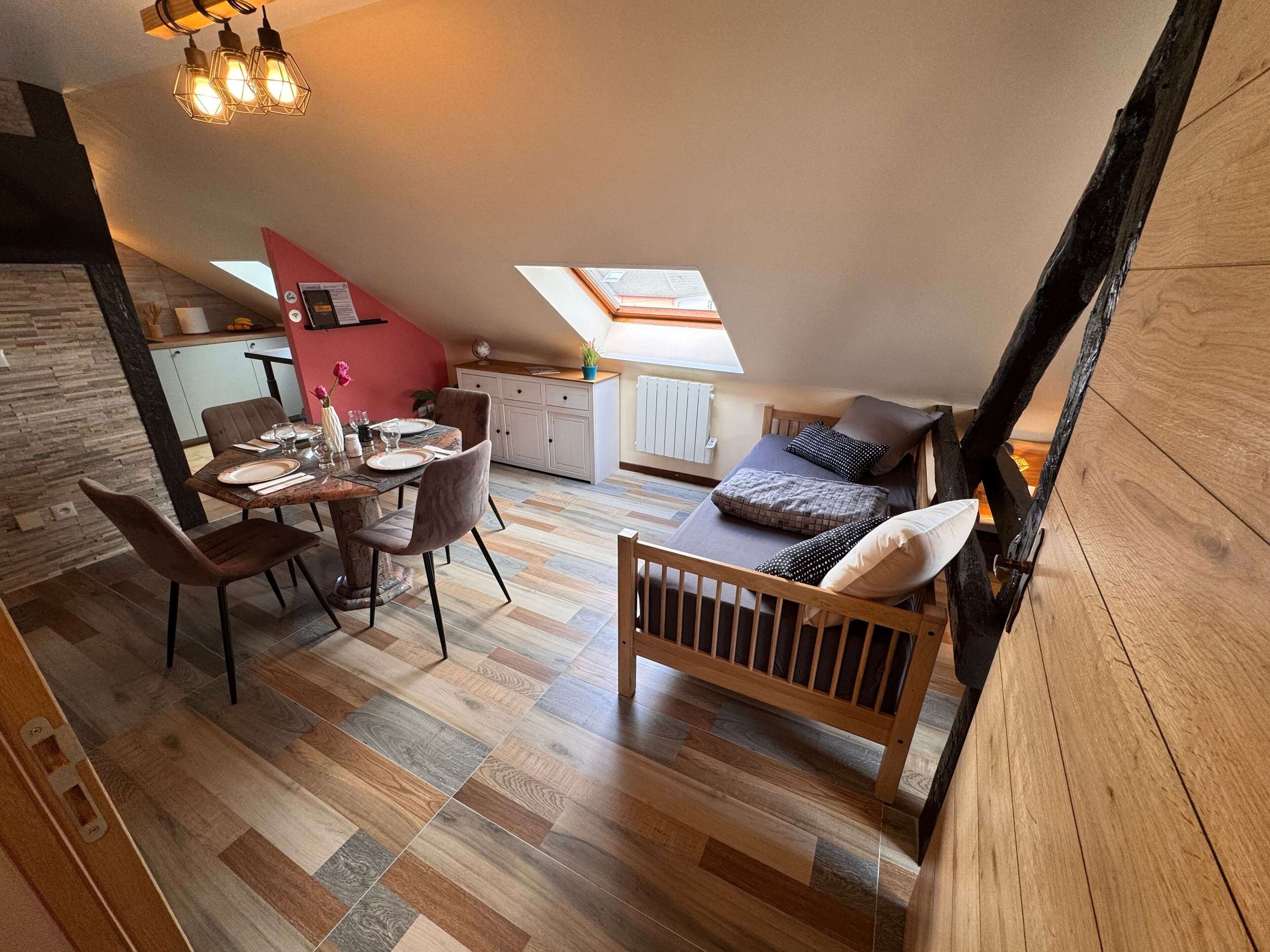 Um quarto no estilo loft, com teto inclinado, área de jantar com mesa e cadeiras de madeira, e uma pequena cozinha compacta.
