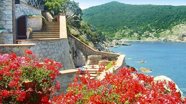 A stone staircase leading to a terrace overlooking a coastal area with a clear view of the sea and lush green hills in the background.