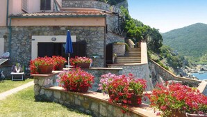 A stone building with a terrace, potted flowers, and a view of the sea and mountains.
