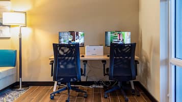 Et hotelværelse med to kontorstole, et skrivebord med en bærbar computer og to skærme, der viser indhold.