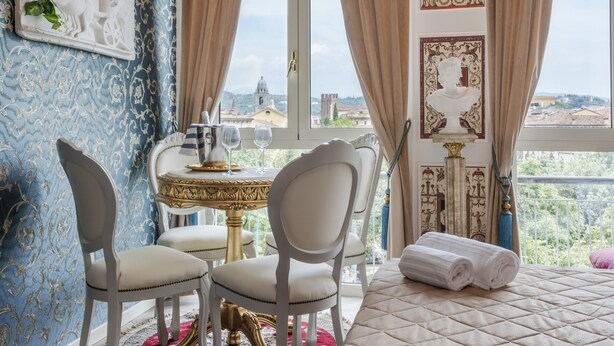 A room with a view of a cityscape, a table with a vase, and a bed with a rolled towel. - Olympus Verona Relais (Verona)