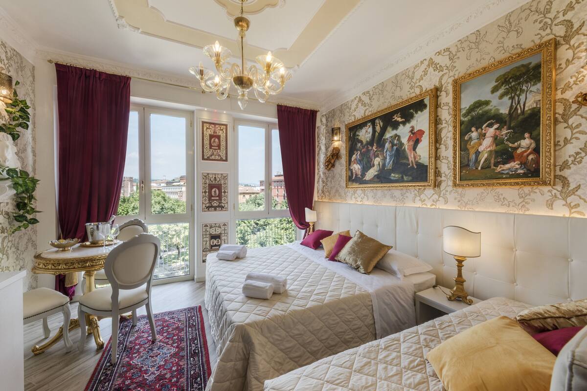 A hotel room with two beds, a chandelier, a dining area with a table and chairs, and a balcony with a view.