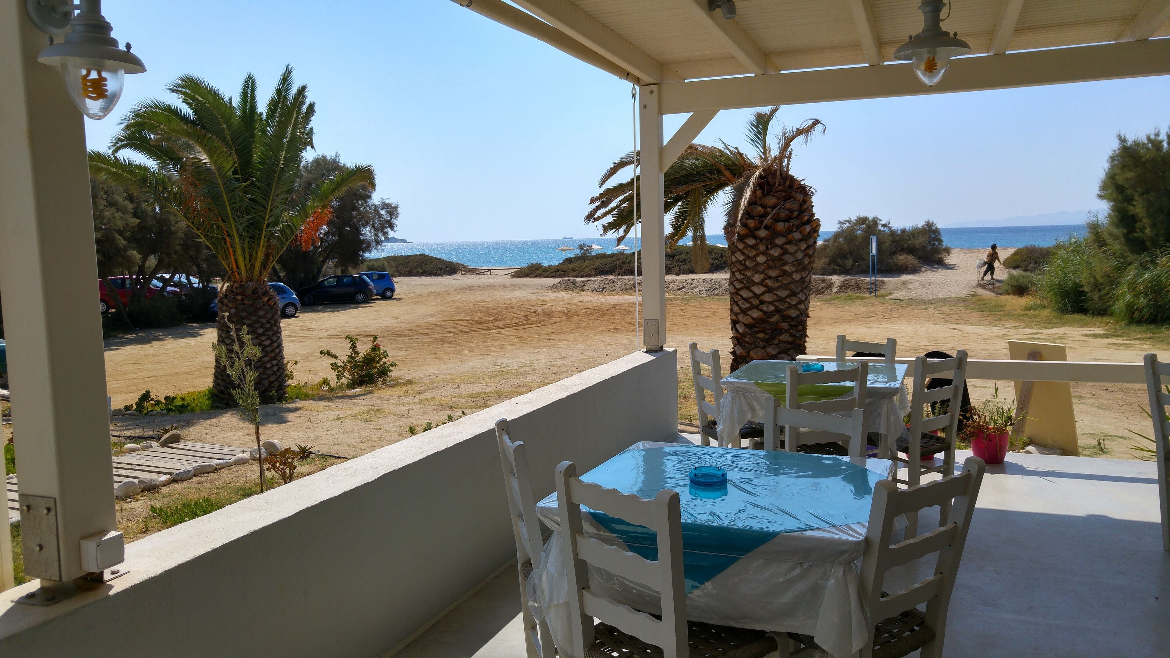 A covered outdoor seating area with a table and chairs, overlooking a beach with palm trees and parked cars.