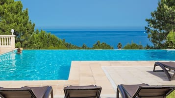 Una piscina con sillones de descanso, vista despejada al océano y zona de terraza con baldosas.