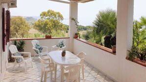 Eine überdachte Terrasse mit weißem Tisch und Stühlen, umgeben von Topfpflanzen und Blick auf die Landschaft.