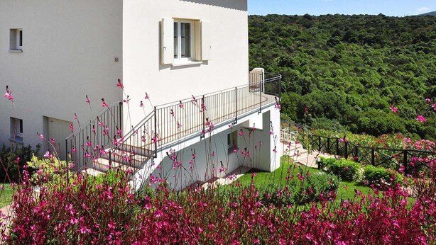 A two-story building with a balcony, surrounded by lush greenery and flowering plants. - Apartment in Golfo Aranci with terrace (Baja Sardinia (SS))