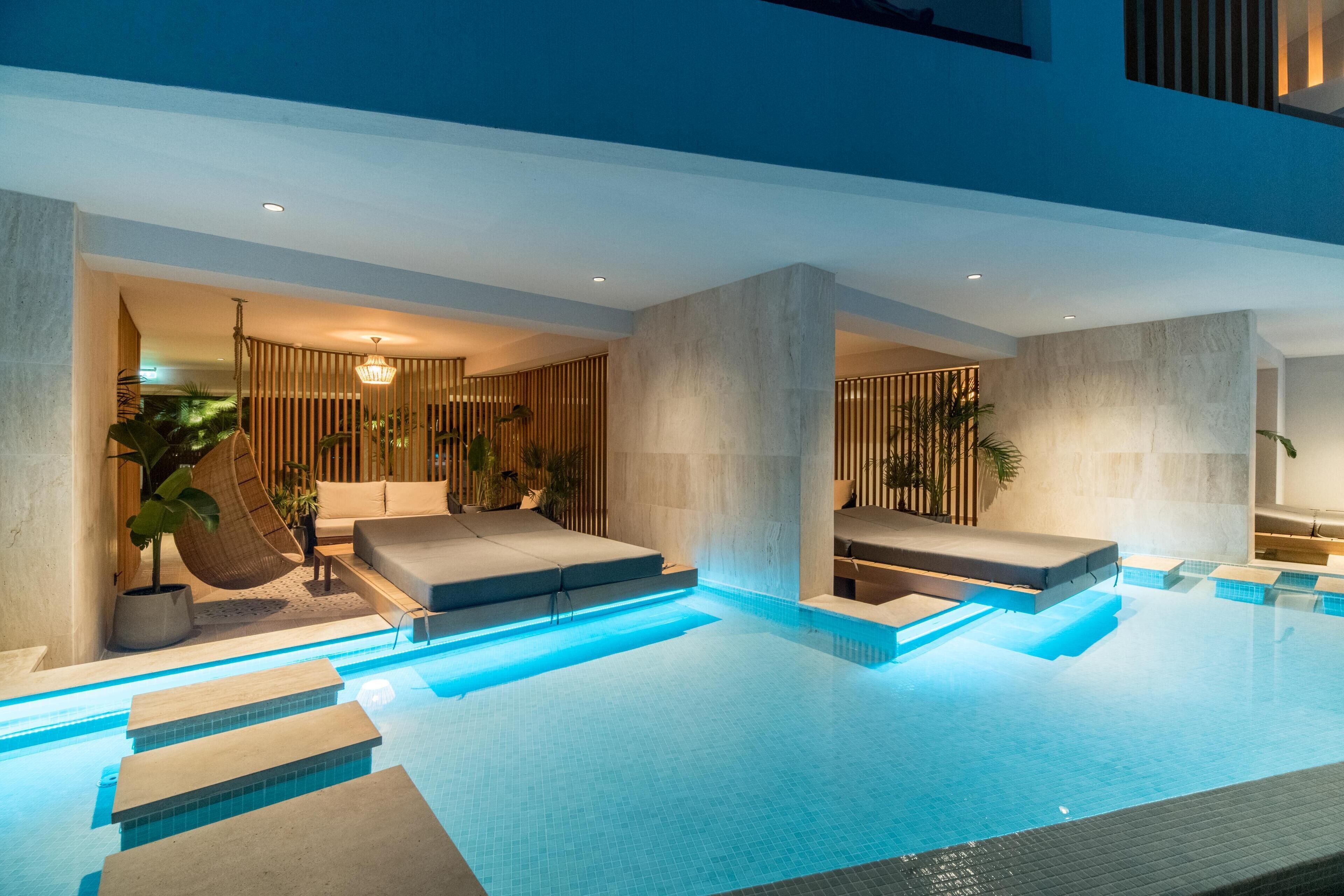 A modern indoor swimming pool with clear blue water, stone steps leading into the pool, and a lounge area with seating and plants.