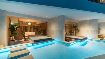 A modern indoor swimming pool with clear blue water, stone steps leading into the pool, and a lounge area with seating and plants.