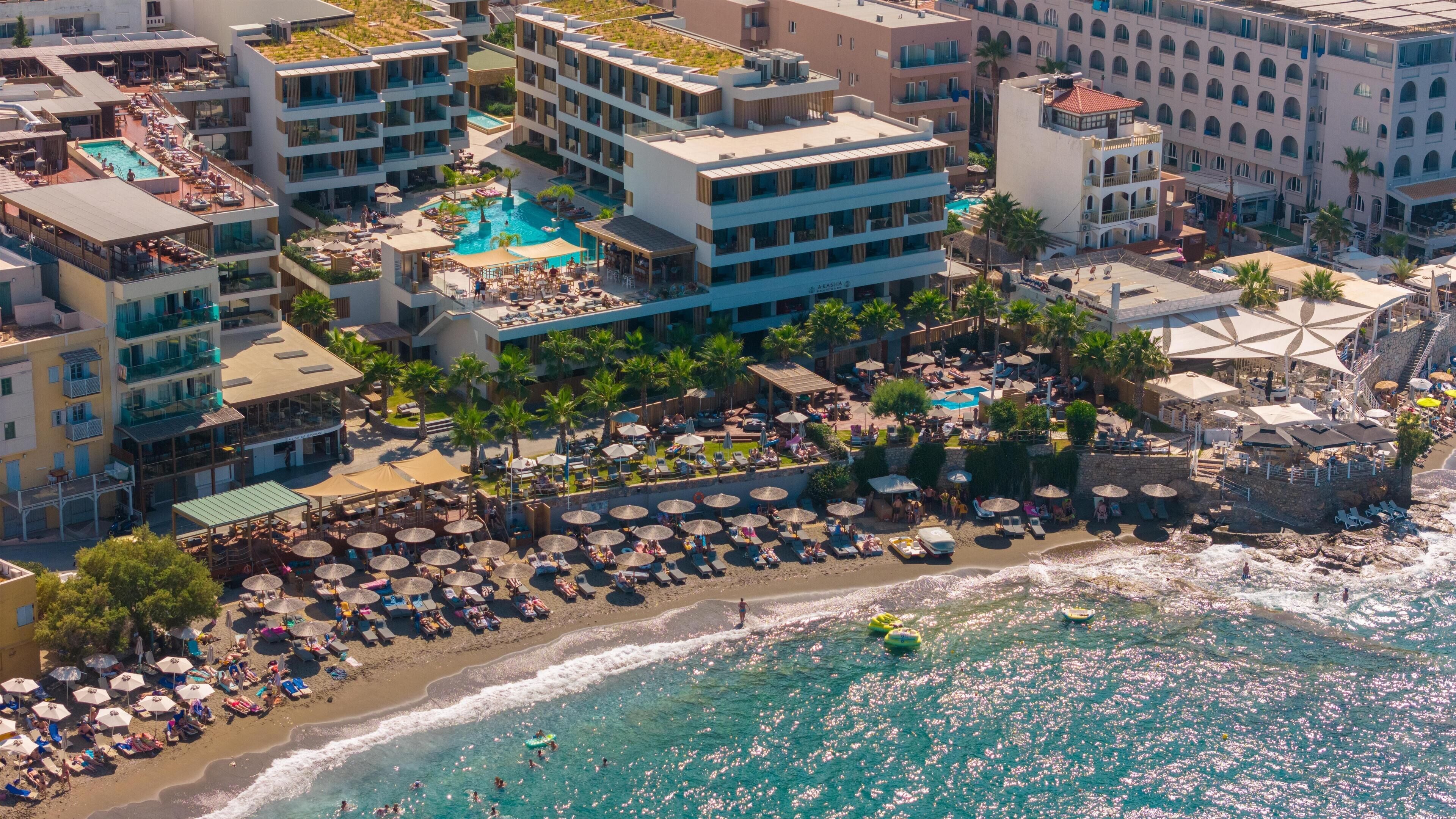 Vista aérea de un complejo turístico costero con varios edificios, una piscina central y una zona de playa con tumbonas y sombrillas.