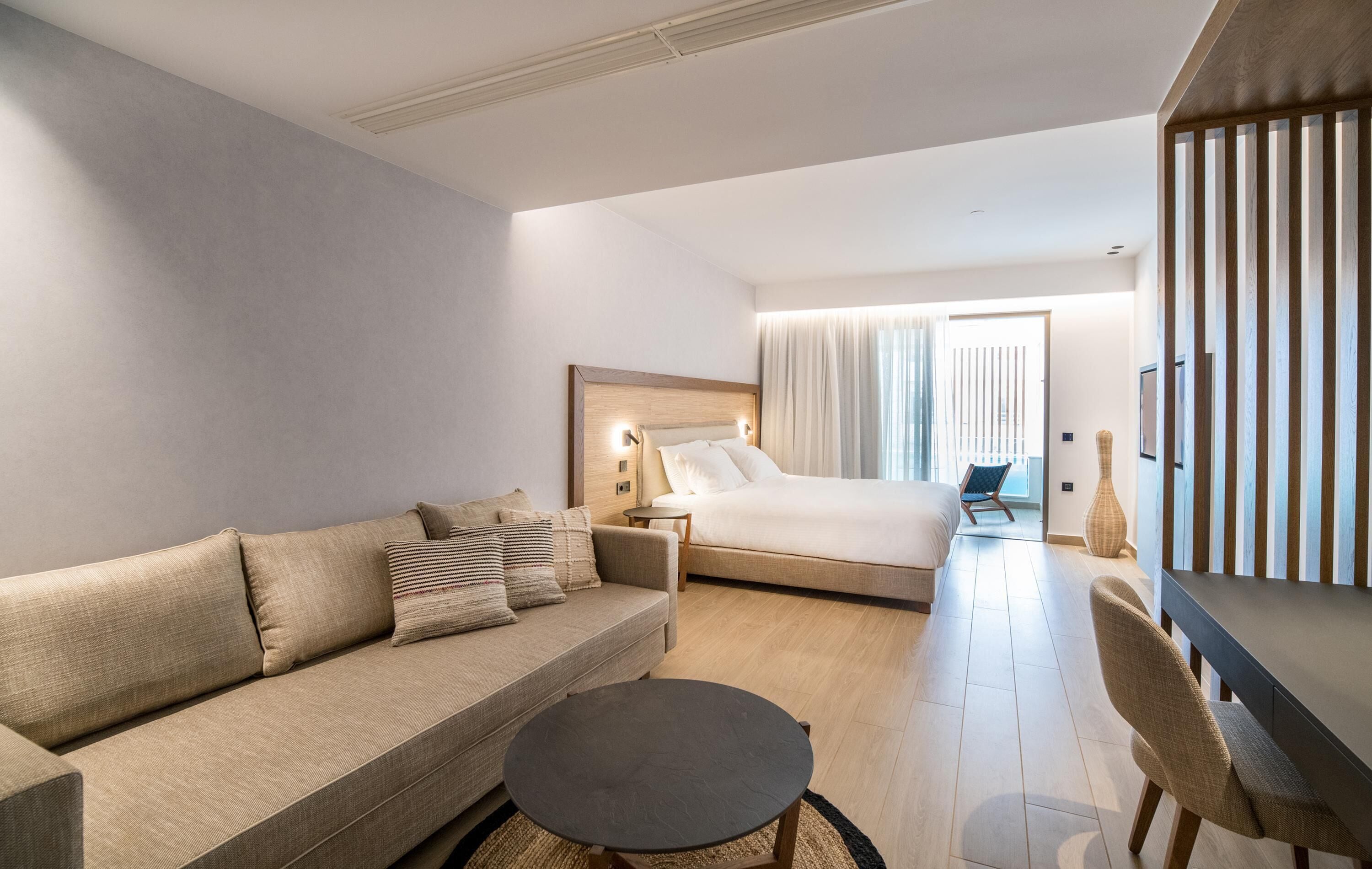 A modern hotel room with a sofa, a round coffee table, a bed with a wooden headboard, a nightstand, and a large window with curtains.