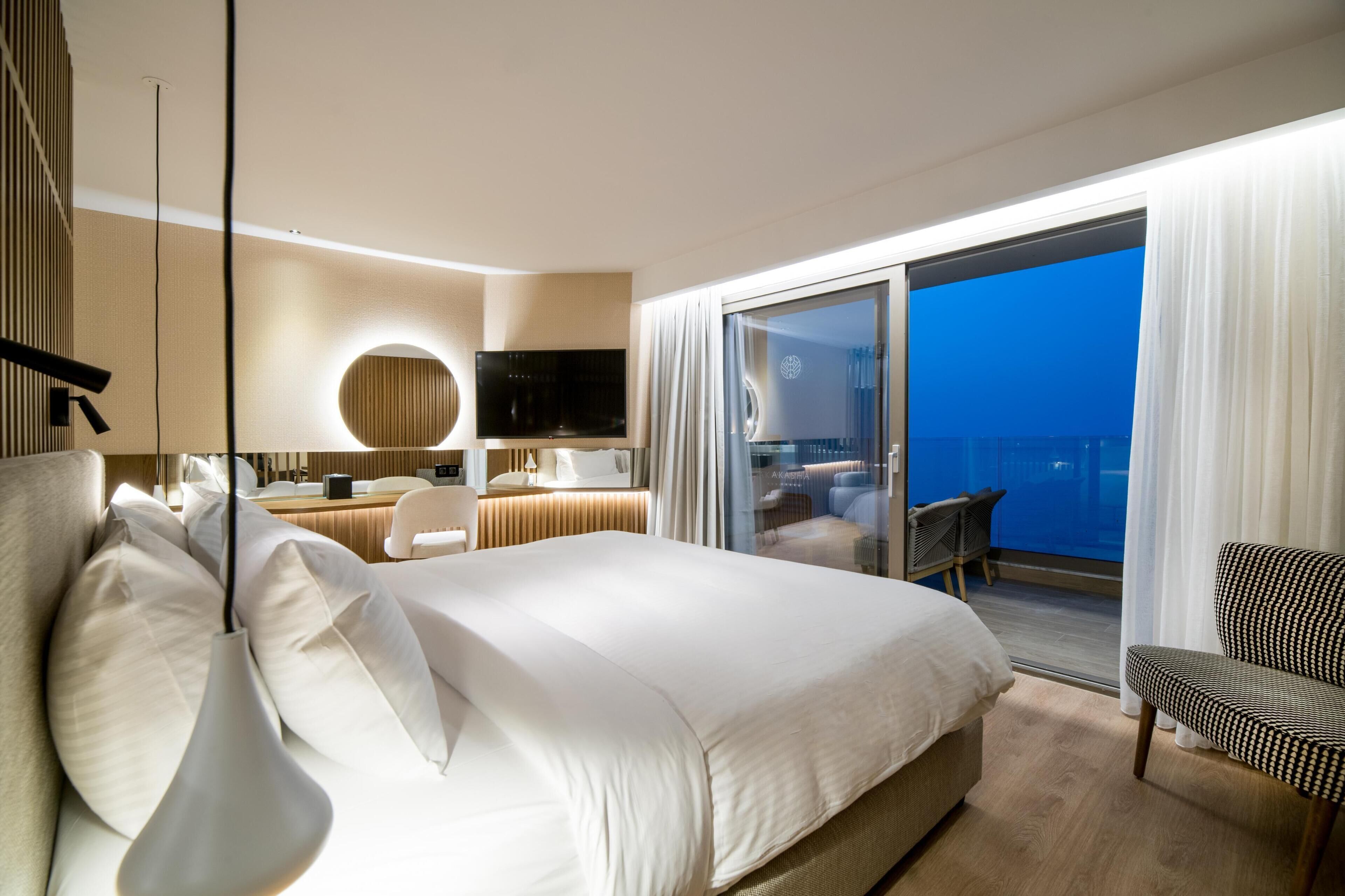 A modern hotel room with a large bed, a desk, a chair, and a balcony with a view.