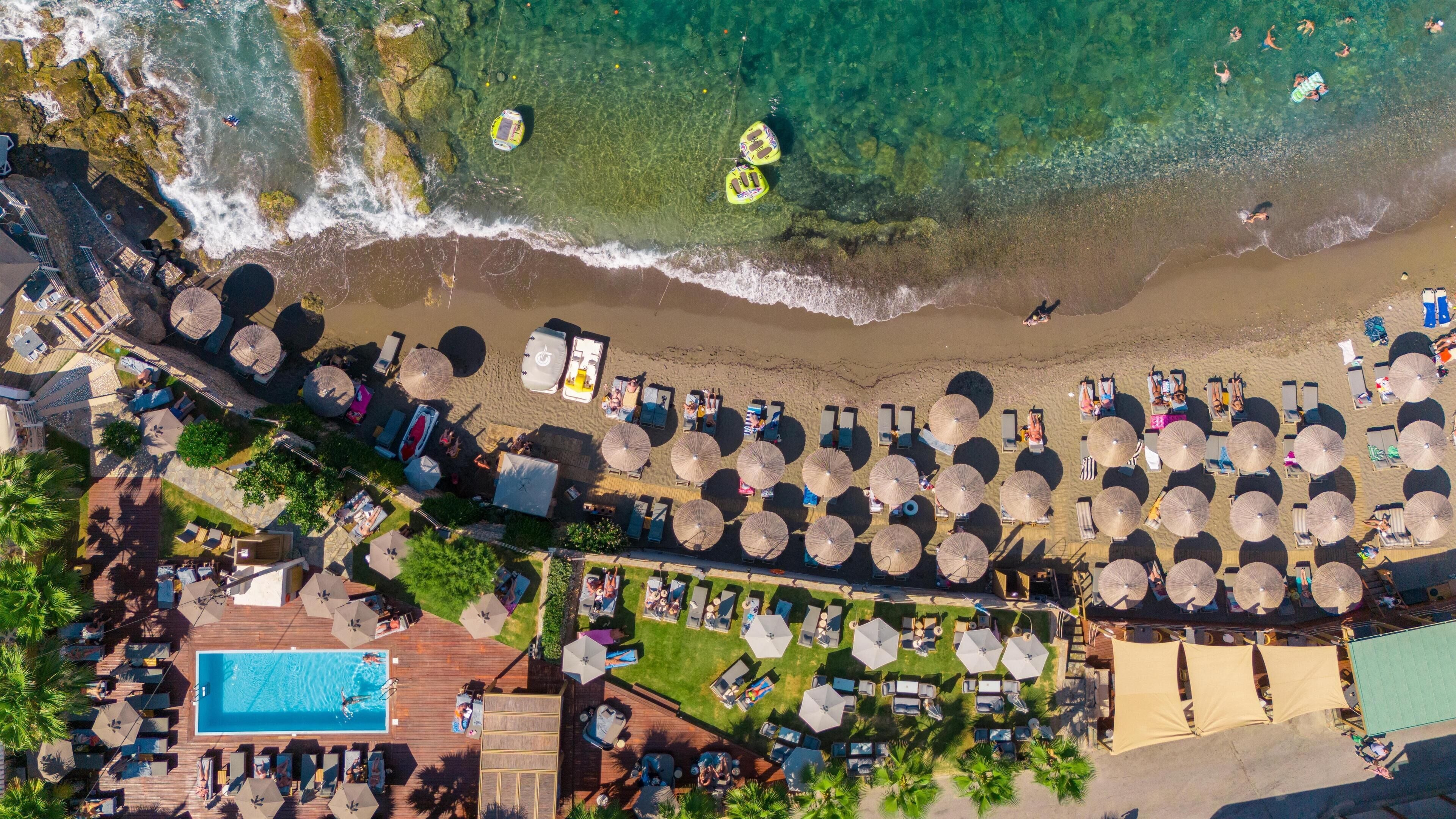 An aerial view of a beach resort with sun loungers, umbrellas, a swimming pool, and a beach area with people and boats.