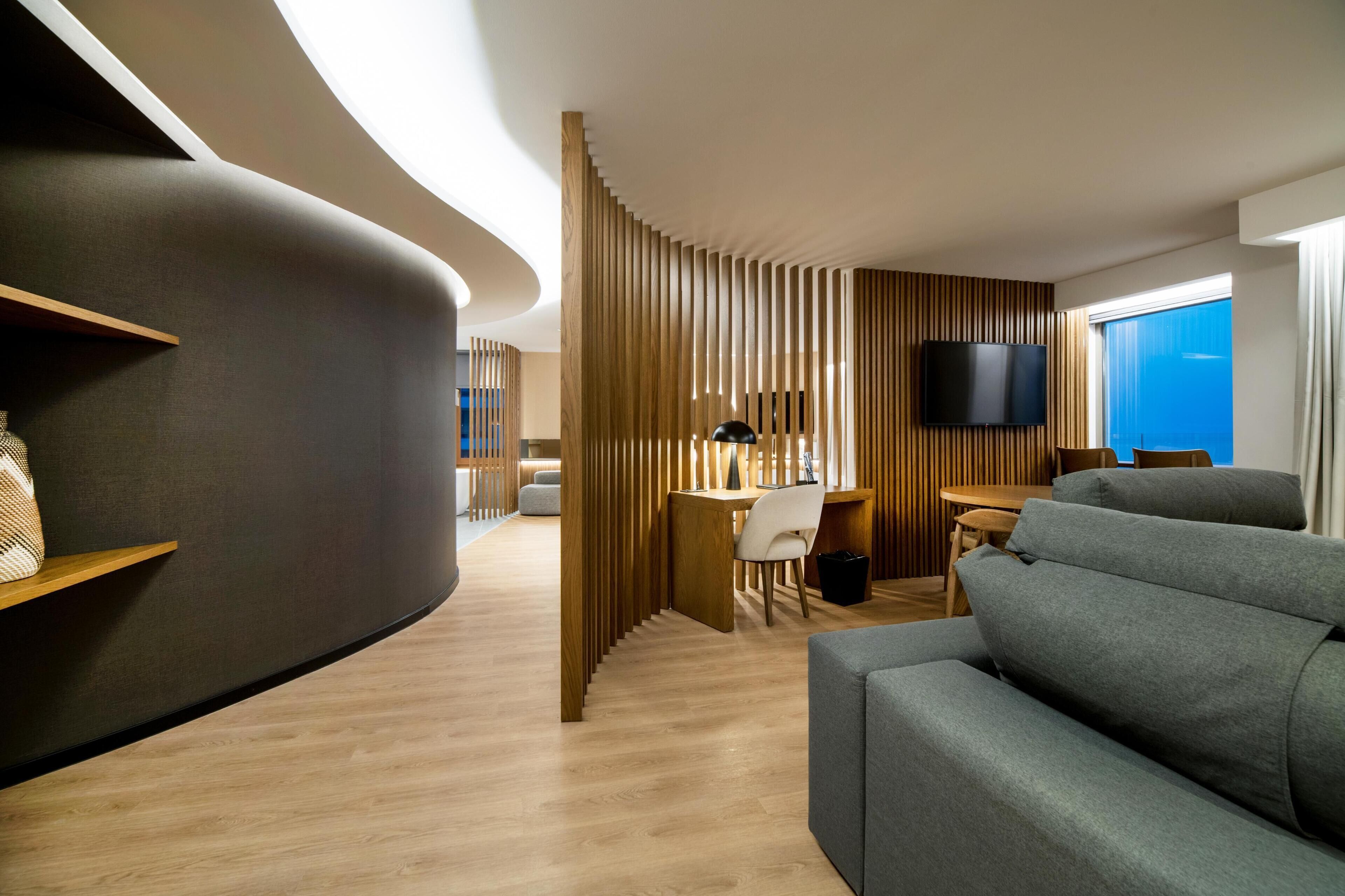 A modern living room with a curved wall, a grey sofa, a wooden dining table with chairs, and a TV mounted on the wall.