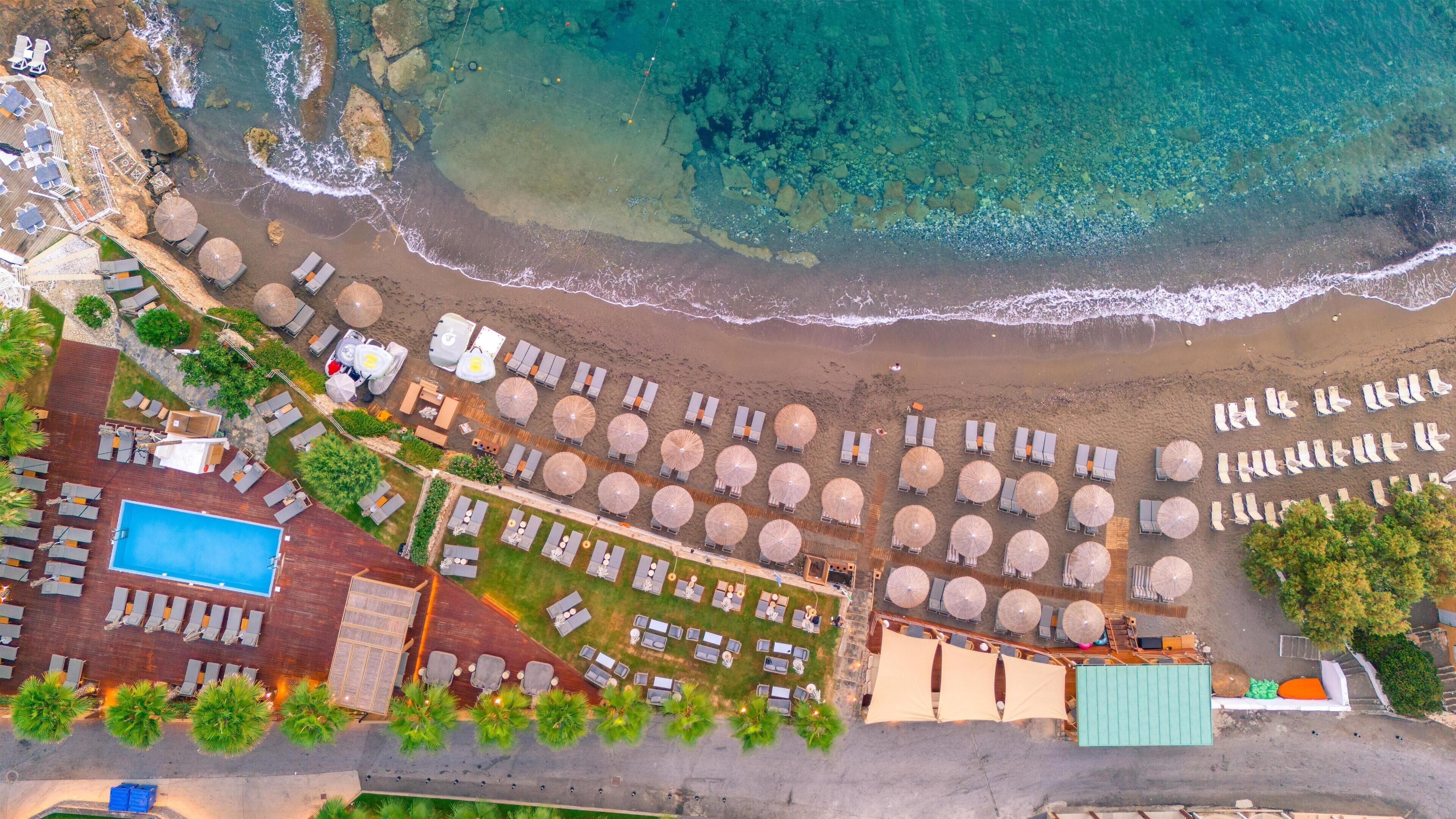 An aerial view of a beach resort with rows of sun loungers and umbrellas, a swimming pool, and a building with a pool deck.