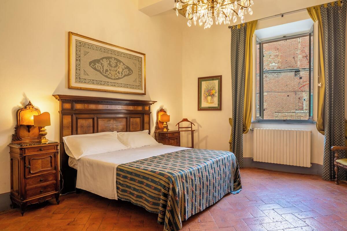 Une chambre à coucher bien rangée, avec un lit en bois, deux tables de chevet avec des lampes, un tableau encadré au-dessus du lit, une fenêtre avec des rideaux et un radiateur.