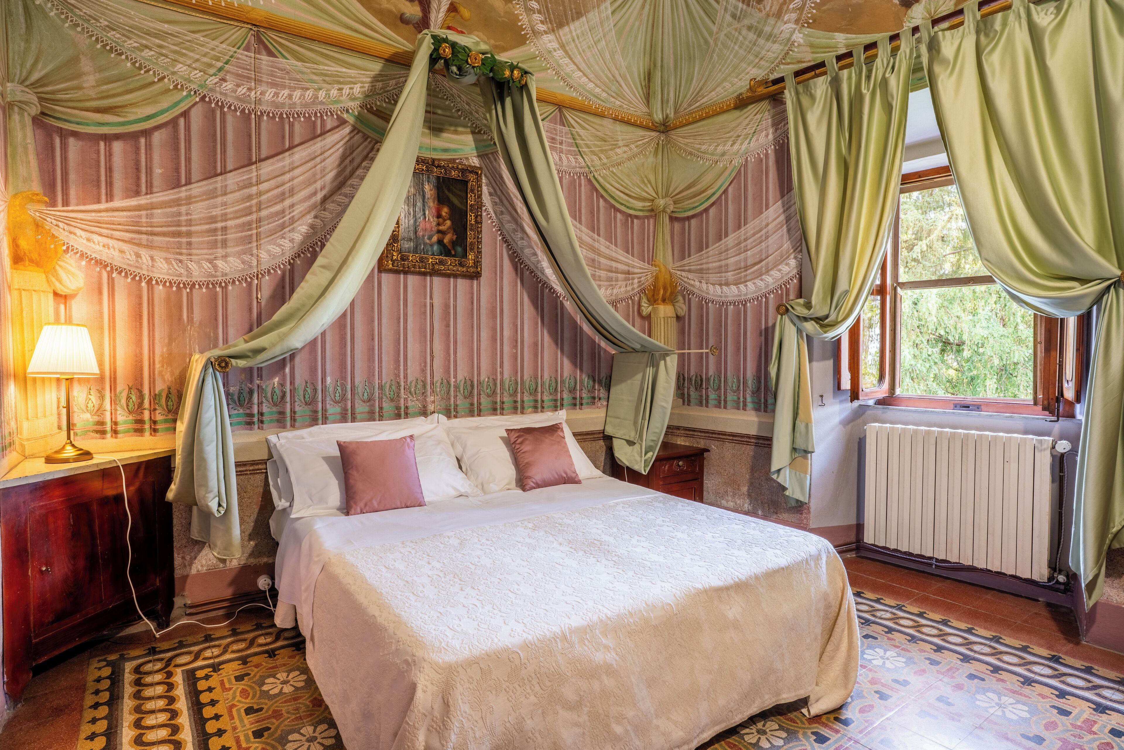 Un dormitorio con cama a dosel, una mesita de noche con lámpara, un radiador y una ventana con cortinas.