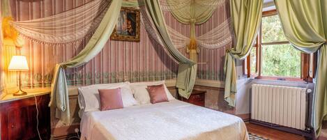 Ein Schlafzimmer mit einem Himmelsbett, einem Nachttisch mit Lampe, einem Heizkörper und einem Fenster mit Vorhängen.